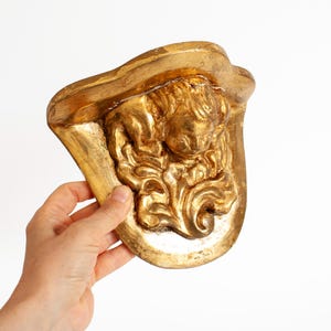 May include: A gold-colored, ornate wall decor piece featuring a sculpted cherub and floral details. The decorative item has a curved shape and is held against a white background. The cherub is the focal point of the design.