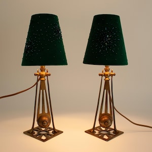 May include: A pair of antique table lamps with dark green velvet shades adorned with dark beads. The lamps have a brass-colored metal base with a geometric design and a square base. The lamps are wired with a brown cord.