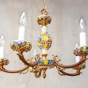 May include: A vintage chandelier with five lights. The fixture features a gold-toned metal frame with white candle holders and colorful floral accents. The bulbs are clear and teardrop-shaped, and the chandelier is suspended from a chain.