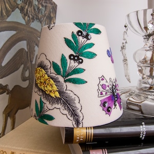 Op de afbeelding: Een witte lampenkap met een bloemen- en vlinderontwerp. De lampenkap is versierd met groene bladeren, gele en zwarte bloemen en paarse en zwarte vlinders. De vlinders hebben iriserende pailletten op hun vleugels.
