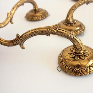 Può includere: Un set di bracci per applique da parete in metallo color oro con design curvi e decorati, caratterizzati da intricati dettagli a forma di foglia. Ogni braccio termina con una base circolare dal bordo smerlato e una piastra piatta per candele.