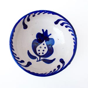 May include: A round ceramic bowl with a white base and hand-painted cobalt blue designs. The interior features a central pomegranate motif with dotted details, surrounded by a decorative border of repeating blue brushstrokes along the rim.