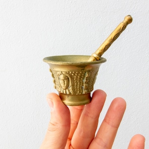 Könnte beinhalten: Ein kleiner, goldfarbener Mörser und Stößel mit dekorativen Details. Der Mörser hat einen runden Boden und eine breite, flache Schale. Der Stößel ist ein langer, dünner Stab mit abgerundetem Ende.