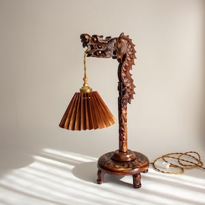 Puede incluir: Lámpara de mesa de madera tallada con un diseño de dragón detallado. La lámpara tiene una pantalla plisada marrón y una base decorativa. La cabeza del dragón sostiene la pantalla y la lámpara tiene un cable dorado.