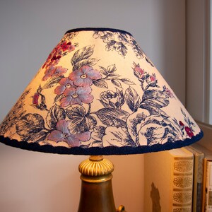 May include: A lampshade with a floral pattern in shades of pink, blue, and black on a cream background. The lampshade has a dark blue trim.