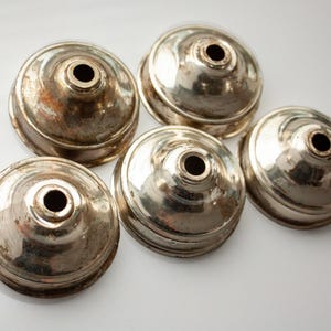 May include: Five silver-toned metal caps with a circular design and a small hole in the centre. The caps are arranged in a semi-circle on a white surface.