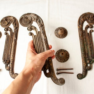 Puede incluir: Tres piezas decorativas de metal adornadas con un diseño de rollo, dos tienen la forma de una 'S' larga y curva y la tercera es una pieza redonda más pequeña. También hay seis pequeños tornillos de metal.