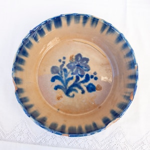 May include: A vintage ceramic plate with a blue and white floral design. The plate has a scalloped edge and is decorated with a stylized floral pattern in shades of blue on a beige background.