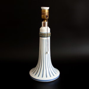 May include: A white ceramic table lamp with a fluted design and blue accents. The lamp has a brass-colored socket and a pull chain. The base is a flared shape with blue stripes and a blue border. The lamp is approximately 12 inches tall.