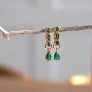 May include: A pair of gold earrings with green gemstone accents. The earrings feature a teardrop-shaped gemstone dangling from a row of smaller gemstones.