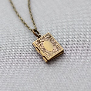 May include: A brass necklace with a small, square locket pendant. The pendant has an oval design in the centre and hangs from a delicate chain. The necklace is set against a soft, textured background. The locket is approximately 1.5 cm square.