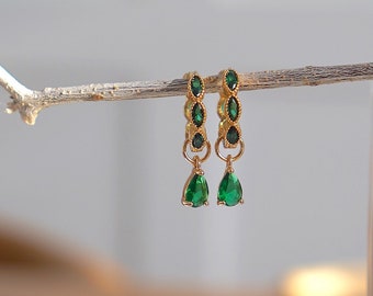 May include: A pair of gold earrings with green gemstone accents. The earrings feature a teardrop-shaped gemstone dangling from a row of smaller gemstones.