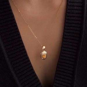 以下が含まれることがあります： 小さな楕円形のペンダントが付いたゴールドカラーのペンダントネックレス。ペンダントは明るい色の中心部とゴールドカラーのフレームです。ネックレスは黒いセーターの上から着用されています。