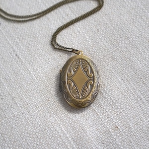 May include: A gold-toned oval locket necklace with an intricate floral design. The locket has a diamond-shaped design in the center.