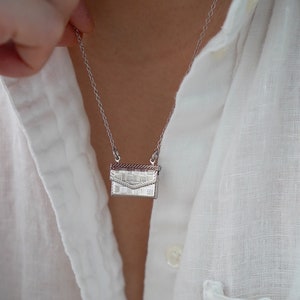 Può includere: Una collana color argento con un piccolo ciondolo a forma di borsa dettagliata. Il ciondolo ha una trama intrecciata e una chiusura a patta. La collana è indossata sopra una camicia bianca abbottonata.