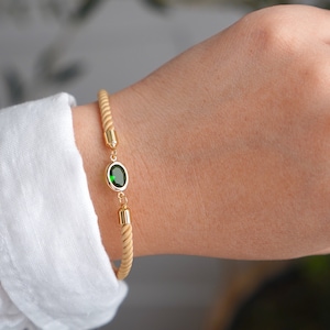 May include: Gold-toned bracelet with a light tan rope-like band and a central oval-shaped green gemstone. The bracelet features gold accents and is worn on a wrist, with a white shirt sleeve visible in the background.