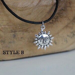 May include: A silver sun pendant necklace with a face on the sun, hanging from a black leather cord. The necklace is on a wooden surface with the text "STYLE B" printed on it.