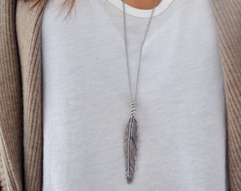 May include: A silver feather pendant necklace hangs over a white v-neck shirt. A tan cardigan is worn over the shirt. A silver watch with a metal band is visible on the wrist.