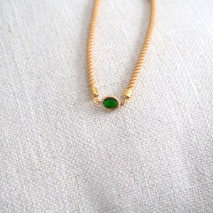 May include: A gold-toned bracelet with a tan rope-like band and a central oval-shaped green gemstone. The bracelet has gold accents at the gemstone and the adjustable clasp. The background is a textured, off-white fabric.