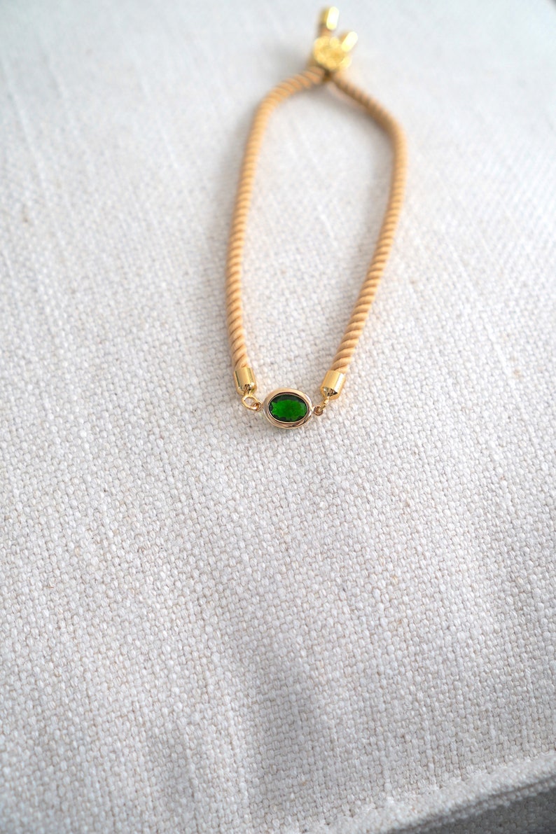 May include: A gold-toned bracelet with a tan rope-like band and a central oval-shaped green gemstone. The bracelet has gold accents at the gemstone and the adjustable clasp. The background is a textured, off-white fabric.