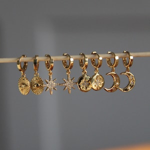May include: A set of eight gold hoop earrings with dangling charms. The charms include a sun with a face, a star, a moon, and a circle with a star inside. The earrings are all on a white background.