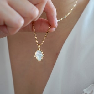 Könnte beinhalten: Eine goldene Kettenkette mit einem weißen Perlmutt-Hamsa-Hand-Anhänger.