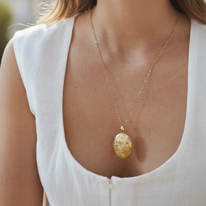 May include: A gold-coloured oval locket pendant necklace. The pendant has an ornate floral design and hangs from a delicate chain. The necklace is worn over a white sleeveless top.