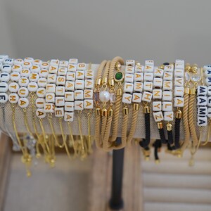 May include: A display of personalized bracelets with white and gold letter beads spelling out names and words. The bracelets are on a neutral-colored display stand, showcasing various designs and styles.
