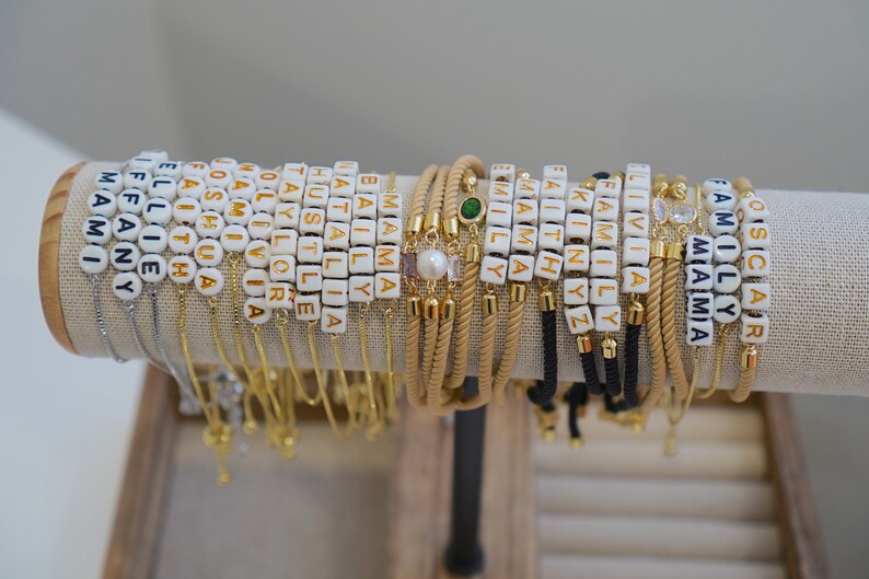 May include: A display of personalized bracelets with white and gold letter beads spelling out names and words. The bracelets are on a neutral-colored display stand, showcasing various designs and styles.