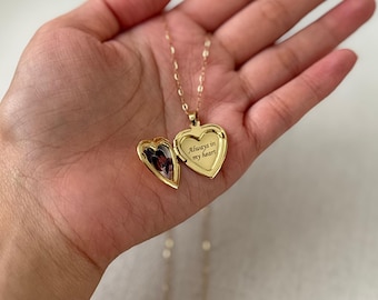 May include: A gold-toned heart-shaped locket necklace on a delicate chain. The locket is open, revealing a small photograph on the left side and the engraved text "Always in my heart" on the right side. The metal has a polished, reflective finish.