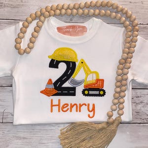 May include: A white short-sleeved t-shirt featuring an embroidered design of a black number two shaped like a road, a yellow construction helmet, an orange and yellow excavator, and an orange traffic cone. The name Henry is embroidered in orange.
