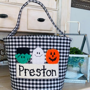 May include: Black and white checkered Halloween trick-or-treat bucket with a green Frankenstein, white ghost, and orange pumpkin applique. The bucket has a white fabric patch with the name "Preston" embroidered on it.