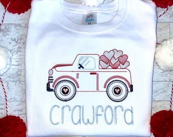 May include: White long-sleeve shirt with a red truck carrying pink and red hearts. The truck has a blue window and the name "Crawford" is embroidered in blue below the truck.