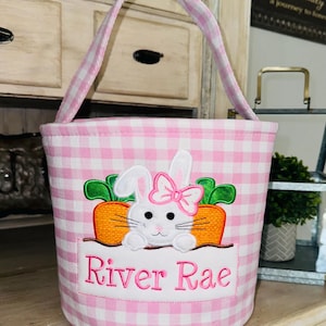 May include: Pink and white gingham Easter basket with a bunny and carrot embroidery. The basket has a handle and says "River Rae" on the front.