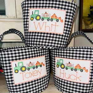 May include: Three fabric baskets featuring a black and white gingham pattern. Each basket has a white rectangular patch embroidered with a green tractor pulling a wagon of orange pumpkins and the names Whit, Toxey, or Huck in orange thread.