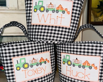 May include: Three fabric baskets featuring a black and white gingham pattern. Each basket has a white rectangular patch embroidered with a green tractor pulling a wagon of orange pumpkins and the names Whit, Toxey, or Huck in orange thread.