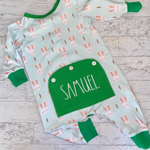 May include: A light blue baby onesie with a green pocket. The onesie has a pattern of white bunnies and orange carrots. The pocket has the name "SAMUEL" embroidered on it.
