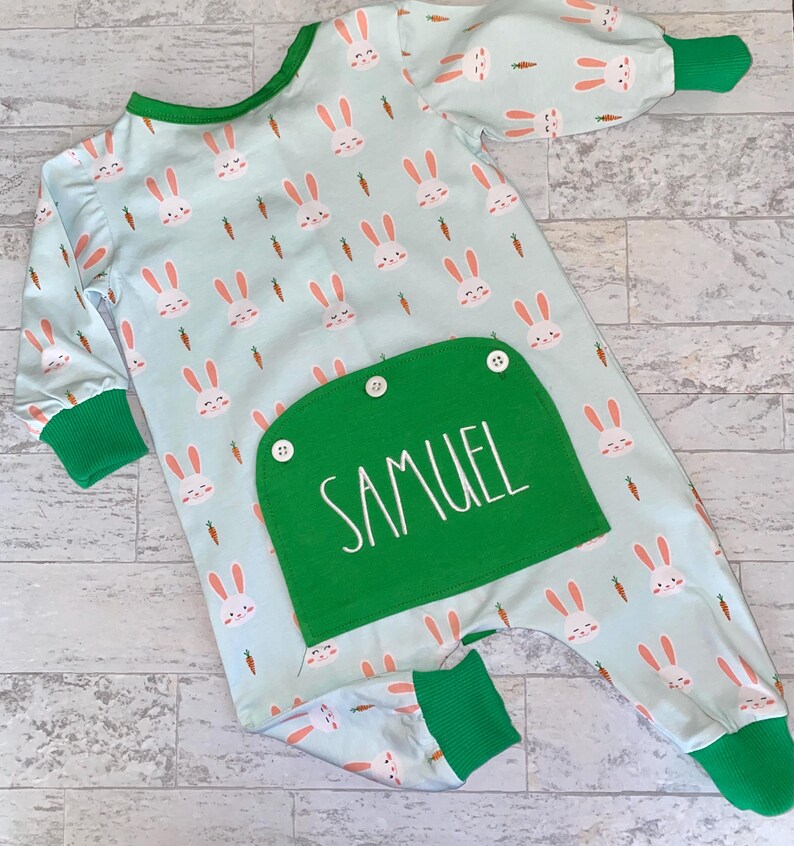 May include: A light blue baby onesie with a green pocket. The onesie has a pattern of white bunnies and orange carrots. The pocket has the name "SAMUEL" embroidered on it.