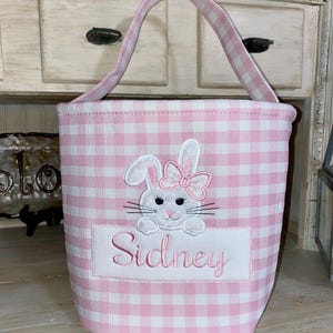 May include: Pink and white gingham Easter basket with a white bunny embroidered on the front. The bunny has a pink bow and the name "Sidney" is embroidered below it.