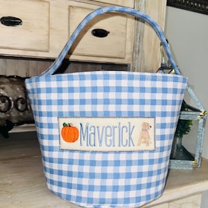 May include: A blue and white gingham fabric tote bag with a handle. The bag features an embroidered patch with the name "Maverick", a pumpkin, and a dog illustration. The bag is sitting on a white surface.
