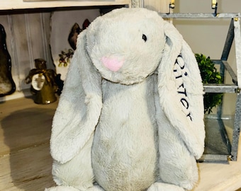May include: A gray plush bunny rabbit with long floppy ears. The bunny has a pink nose and black eyes. The bunny is sitting on a wooden surface. The bunny's ear has the word "Glitter" embroidered on it.