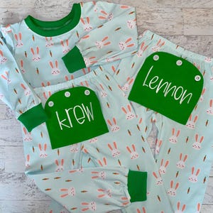 May include: Two pairs of blue pajamas with a bunny and carrot print. The pajamas have green trim and a green pocket with a name on it. One pocket says "Krew" and the other says "Lemon".