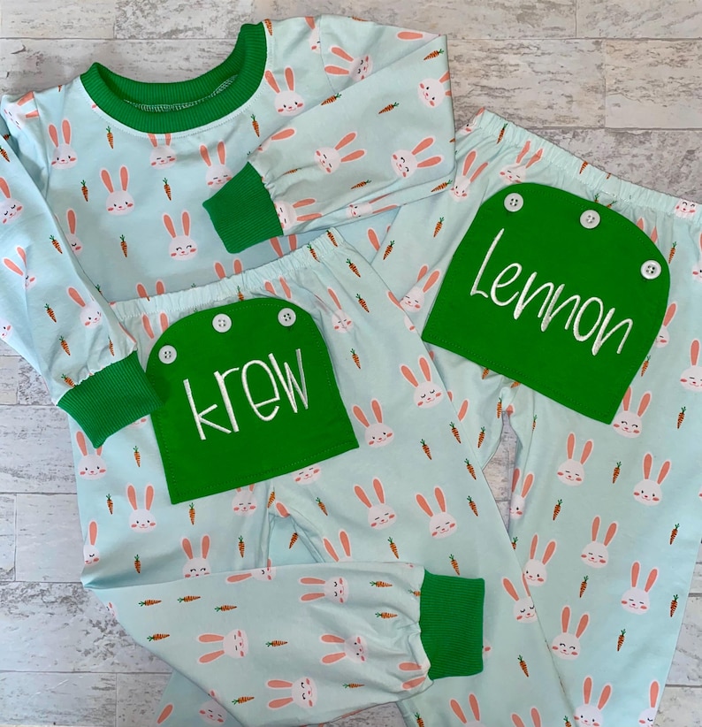 May include: Two pairs of blue pajamas with a bunny and carrot print. The pajamas have green trim and a green pocket with a name on it. One pocket says "Krew" and the other says "Lemon".