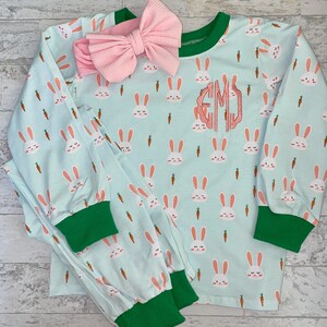 May include: A light blue pajamas set with a bunny and carrot print. The pajamas have green trim and a pink bow headband. The top has a monogrammed "EMJ" on the front.