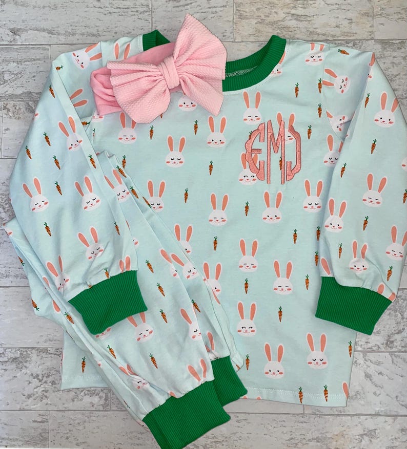 May include: A light blue pajamas set with a bunny and carrot print. The pajamas have green trim and a pink bow headband. The top has a monogrammed "EMJ" on the front.