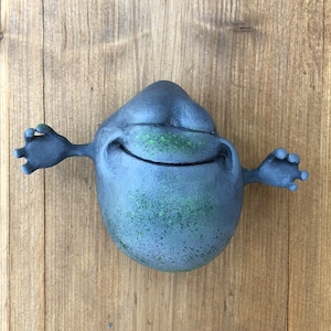 Puede incluir: Una figura gris, sonriente, de dibujos animados con los brazos extendidos. La figura tiene una superficie texturizada con acentos verdes.