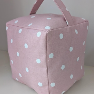 May include: A pink fabric doorstop featuring white polka dots. The cube-shaped doorstop has a matching pink handle on top. A decorative and practical home accessory, it is designed to hold doors open.