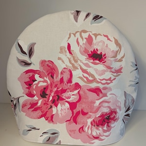 May include: A white fabric kettle cover with a pink and grey floral pattern. The flowers are large and have a textured appearance.