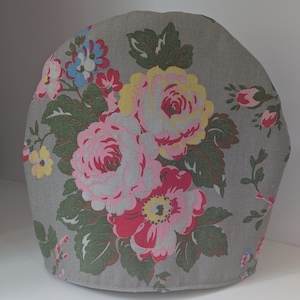 Op de afbeelding: Een grijze theemuts met een bloemenpatroon. Het patroon bevat grote roze, rode en gele rozen met groene bladeren. De muts is gemaakt van stof en heeft een afgeronde vorm.