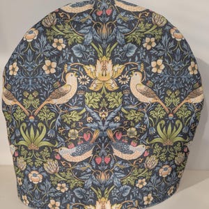 May include: A tea cosy featuring a detailed pattern of birds, flowers, and leaves in beige, green, and yellow on a dark blue background. The fabric tea cosy is designed to keep a teapot warm.